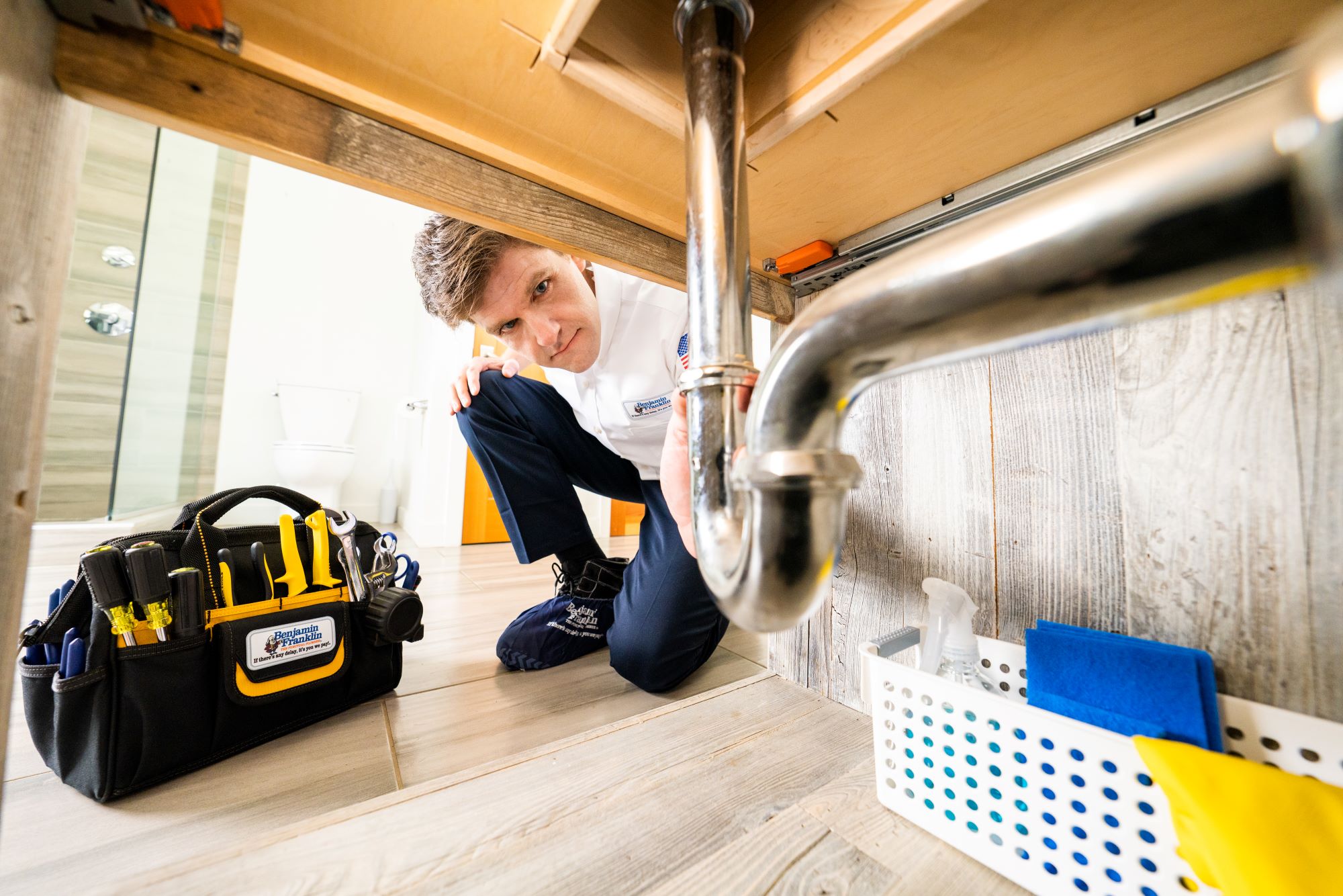 Plumbing business owner in Benjamin Franklin Plumbing uniform looking at a pipe under a sink.