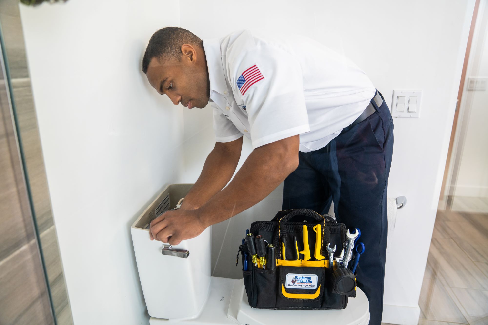 Plumber in a Benjamin Franklin Plumbing uniform with a branded tool bag working on a toilet.