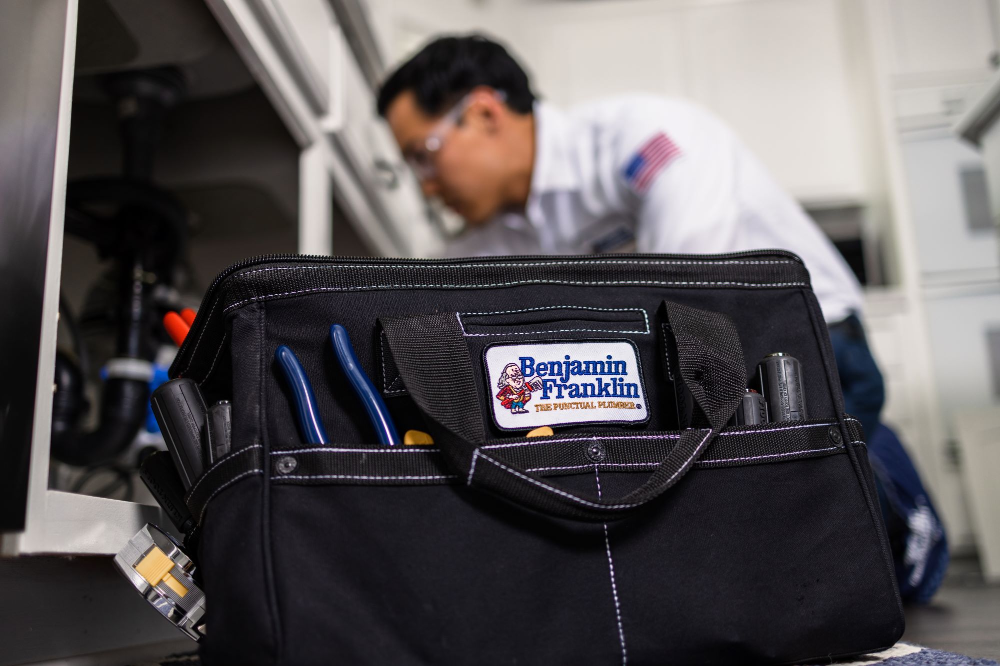 Close up of Benjamin Franklin Plumbing tool bag with plumber working under a sink in the background.